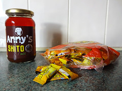 Groceries displayed on a countertop with a white tiled wall behind.  On the left is a glass jar with a label reading “Anny’s Shito”; the only part of the contents visible is a bright red oil that has risen to the top.  On the left is a plastic packet of sweets, each one individually packaged in a yellowish-orange wrapper.