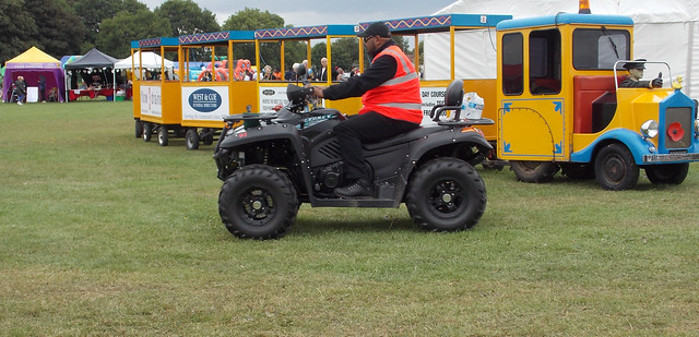 SECURITY OFFICER RIDING ON A QUAD BIKE WITH A FAIRGROUND TRAIN RIDE PASSING IN THE BACKGROUND AT A MUSIC FESTIVAL IN AN EAST LONDON BOROUGH STREET PARK VENUE EVENT ENGLAND DSCN0848