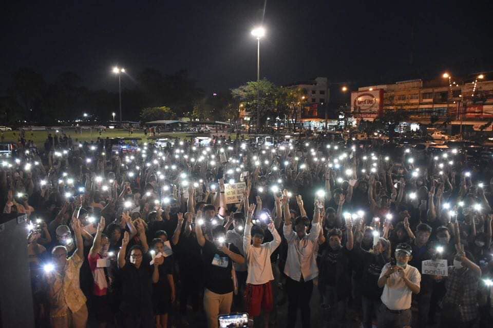A wide shot of the demonstration at Thung Na Choey Park in Chantaburi after dark, showing the protestors holding up their phones with the flashlight turned on. 