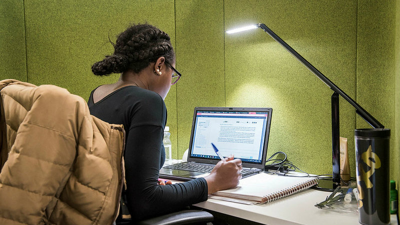 A female, black student sat in front of her laptop writing some notes. There is a bright lit desk lap over her head. The wall in front of her is green.
