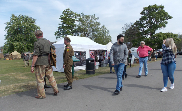 TWO MEN GERMAN MILITARY SOLDIER RE-ENACTORS   WEARING SECOND WORLD WAR UNIFORMS AND WALKING AND ONE STANDING AS A LADY WITH LONG BLONDE HAIR WEARING BLUE DENIM JEANS WALKS PAST AT THE ROYAL GUNPOWDER MILLS VE DAY CELEBRATION EVENT IN AN EAST LONDON BOROUG