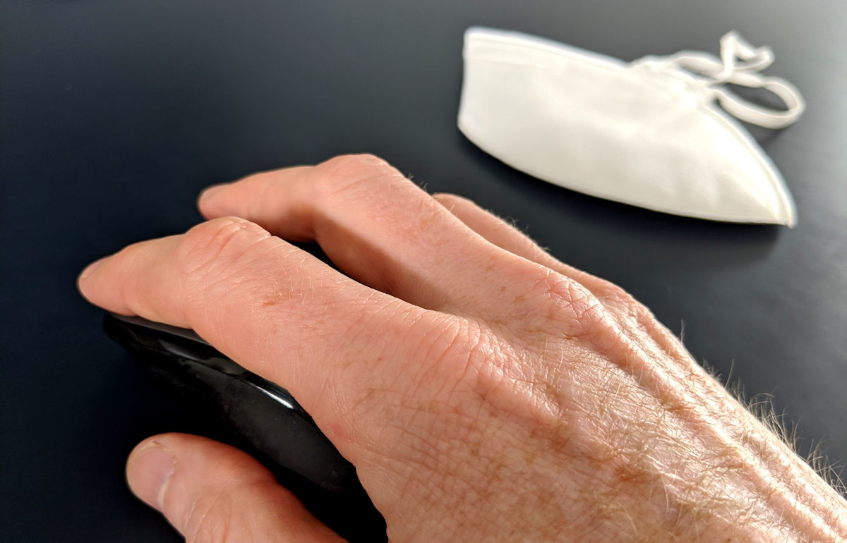A hand on a computer mouse, with a face mask in the background.