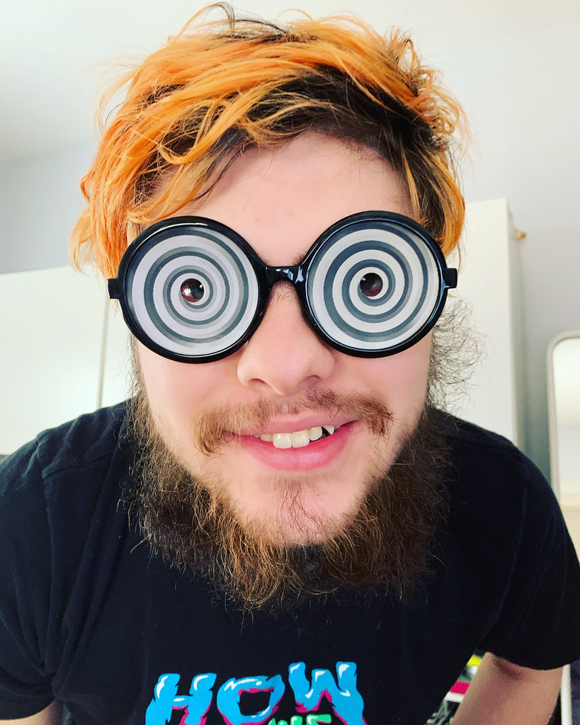 Young man with a scruffy beard and orange hair wearing novelty hypno glasses with black and white swirls