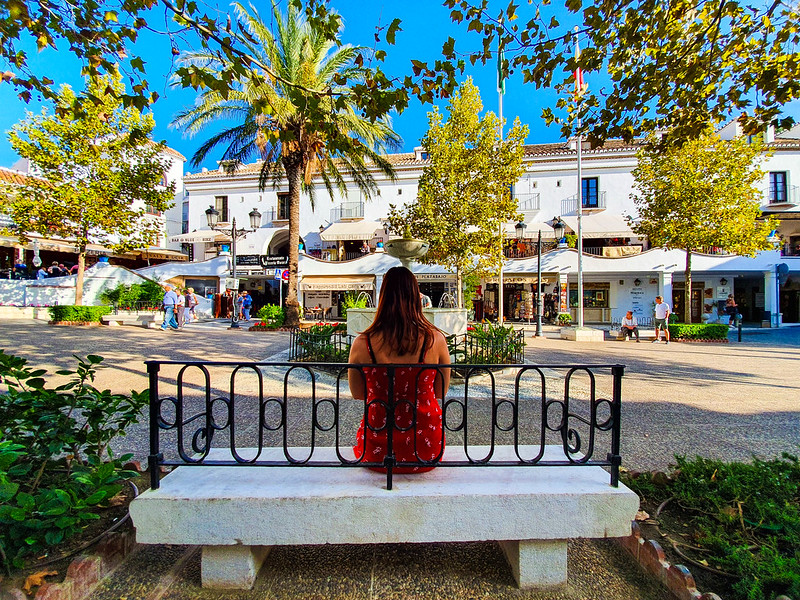 I am sitting on a bench, with my back at the camera. I am wearing a red dress. In front of me there is a small square dotted with palm trees. All the buildings around the square are white, and have shops or cafes at the ground floor.
