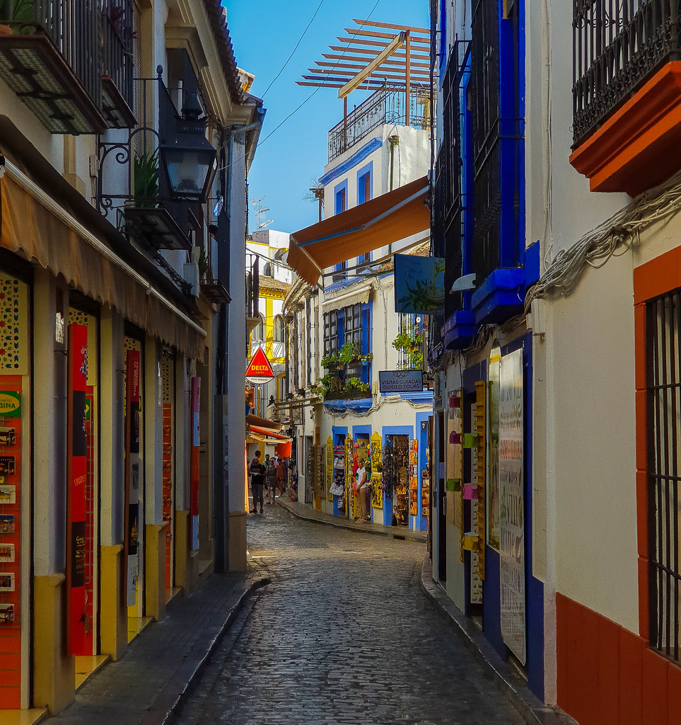 A very narrow and colorful street, with houses painted in different colors around their doors and windows. Most of the houses are shops, and have their merchandise hanging outside, such as fridge magnets, bags, clothes.
