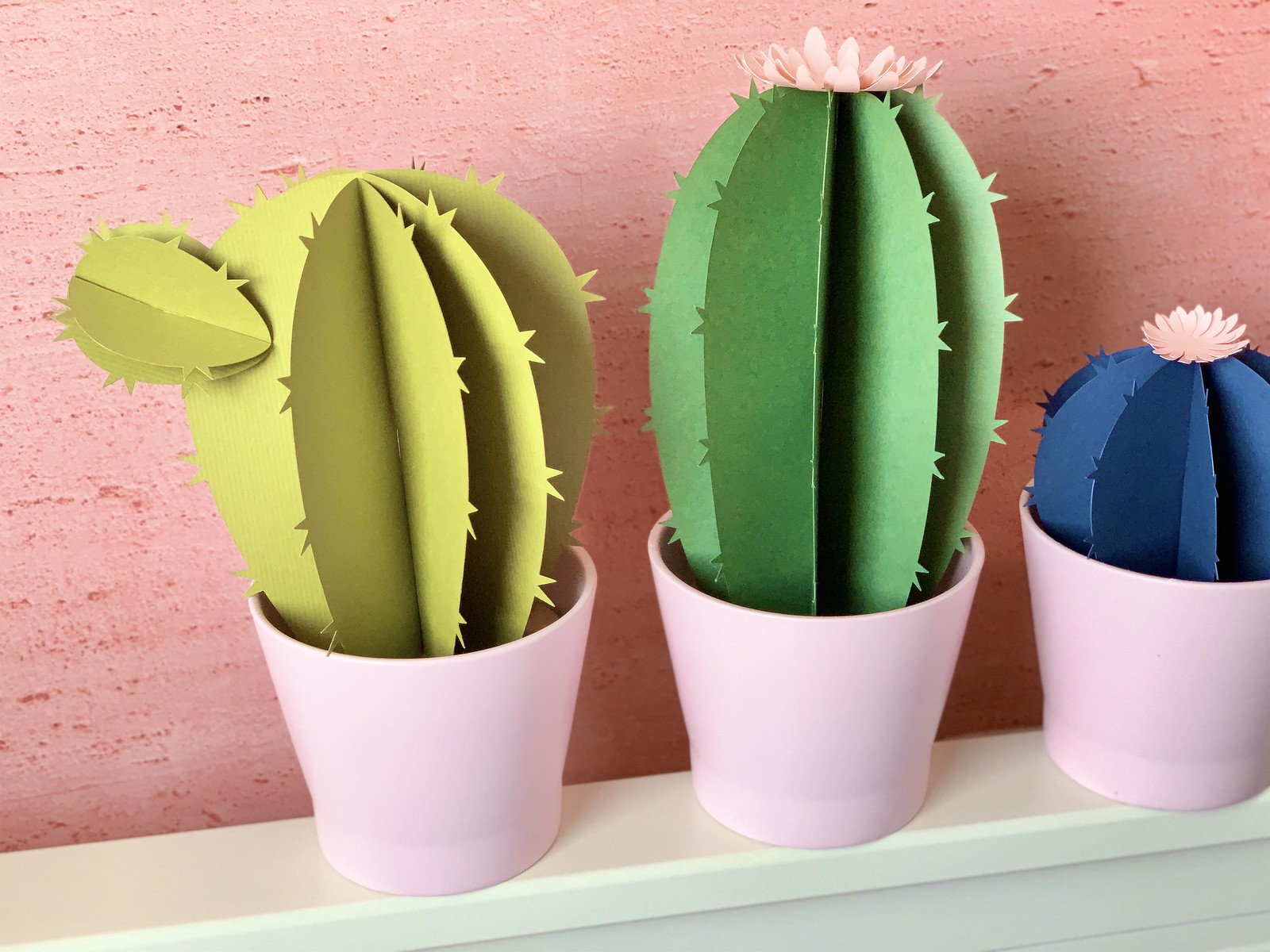 Close up of three paper cactuses. One pale green, one dark green, one dark blue, against a pink wall.