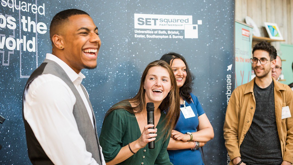 Four young people smile and laugh, standing in front of a SETsquared Partnership backdrop at an event.
