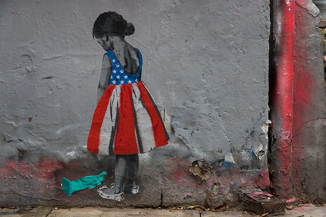 Street art of little girl wearing American Flag dress looking down at a broken Statue of Liberty
