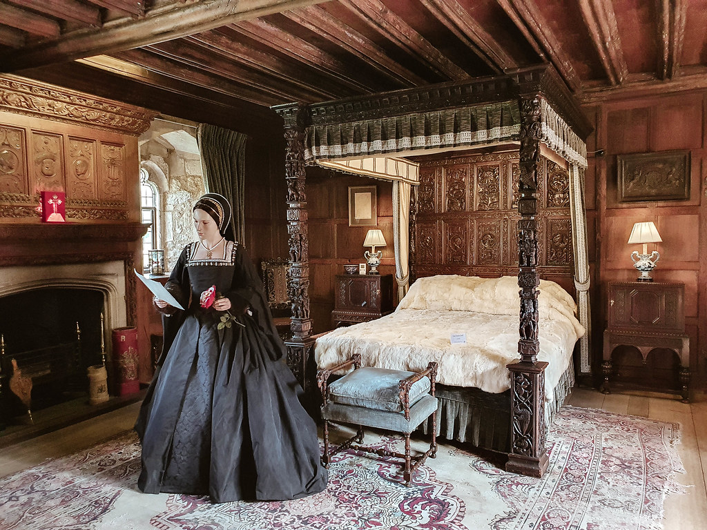 A photo from the oldest room inside Hever Castle, with the original wooden beams on the ceiling. In the middle of the room there is a wooden four poster bed which each of the posts carved with intricate designs. The wood paneling on the wall behind the bed is sculpted as well with different symbols. In front of the bed there is a mannequin representing Anne Boleyn holding a document in her right hand and a red rose in her left one. 