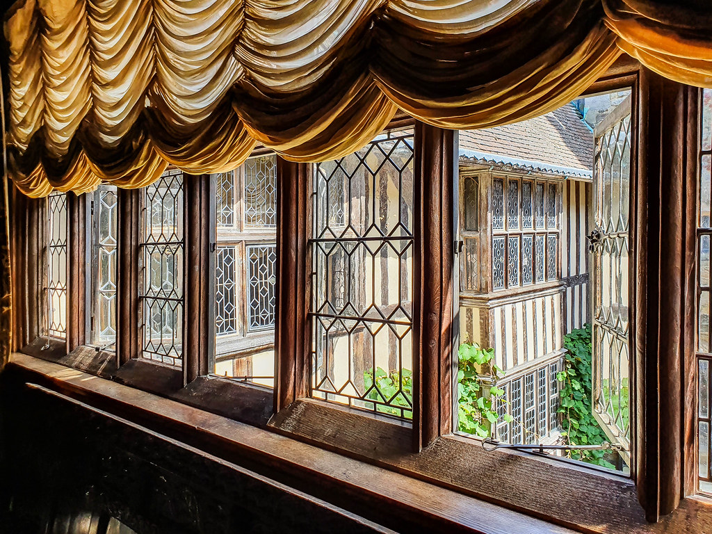 A view of the open windows, seen from the inside of the castle. The windows are Tudor style, with wooden frames and a lattice effect on the glass. Above the windows there is a light brown heavy curtain tied in folds.