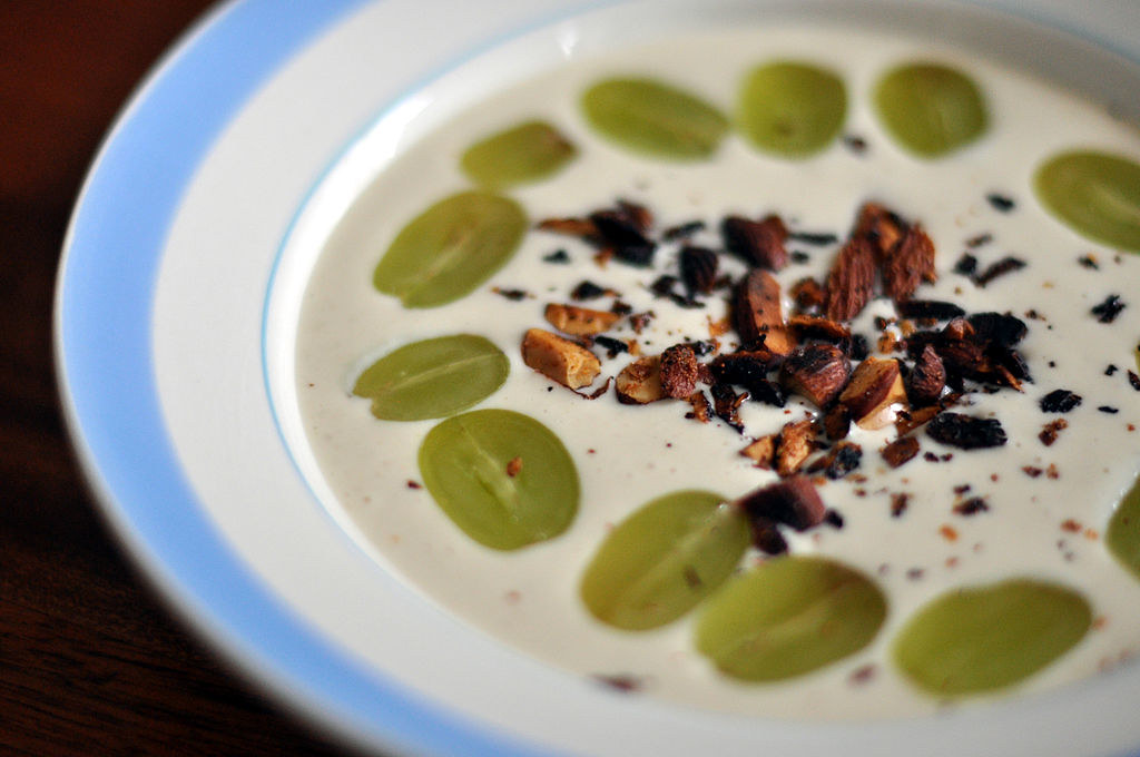 A bowl of white soup topped with sliced of grape that go around in a circle. In the middle, there are crushed almonds. The bowl is white and has a light blue rim