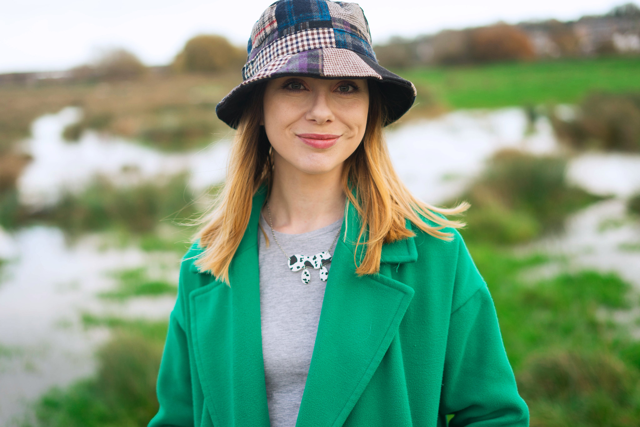 A close up photo of the woman wearing a patchwork tweed bucket hat, a green coat, grey tshirt and a handmade cow print necklace.