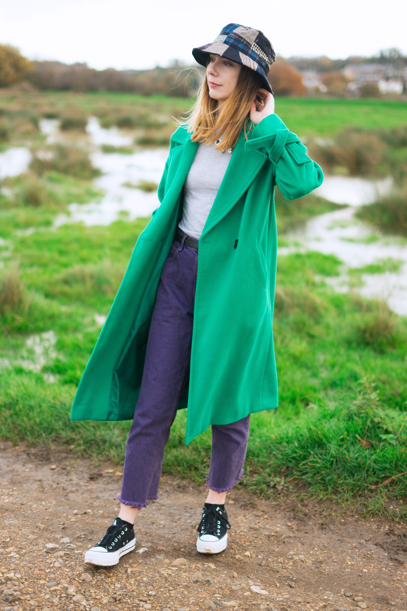 An picture of a white woman wearing a long green coat, a patchwork tweed bucket hat, high waist purple jeans and platform converse outside on a pathway in a field.