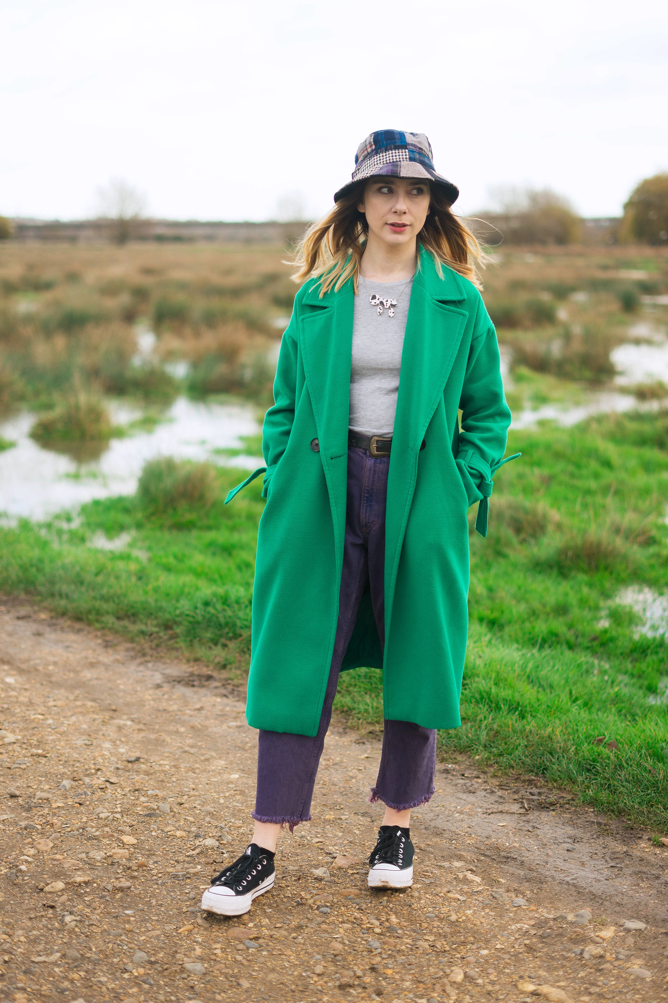 An picture of a white woman wearing a long green coat, a patchwork tweed bucket hat, high waist purple jeans and platform converse outside on a pathway in a field.