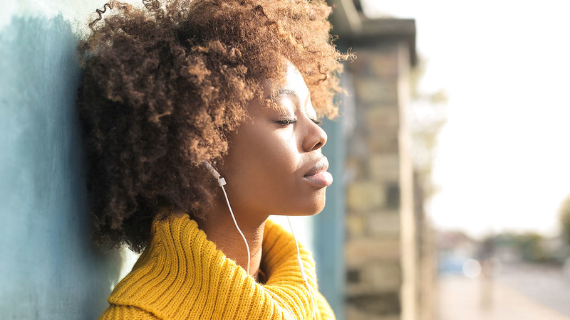 A young woman leaning against a wall listing to music on headphones looking relaxed