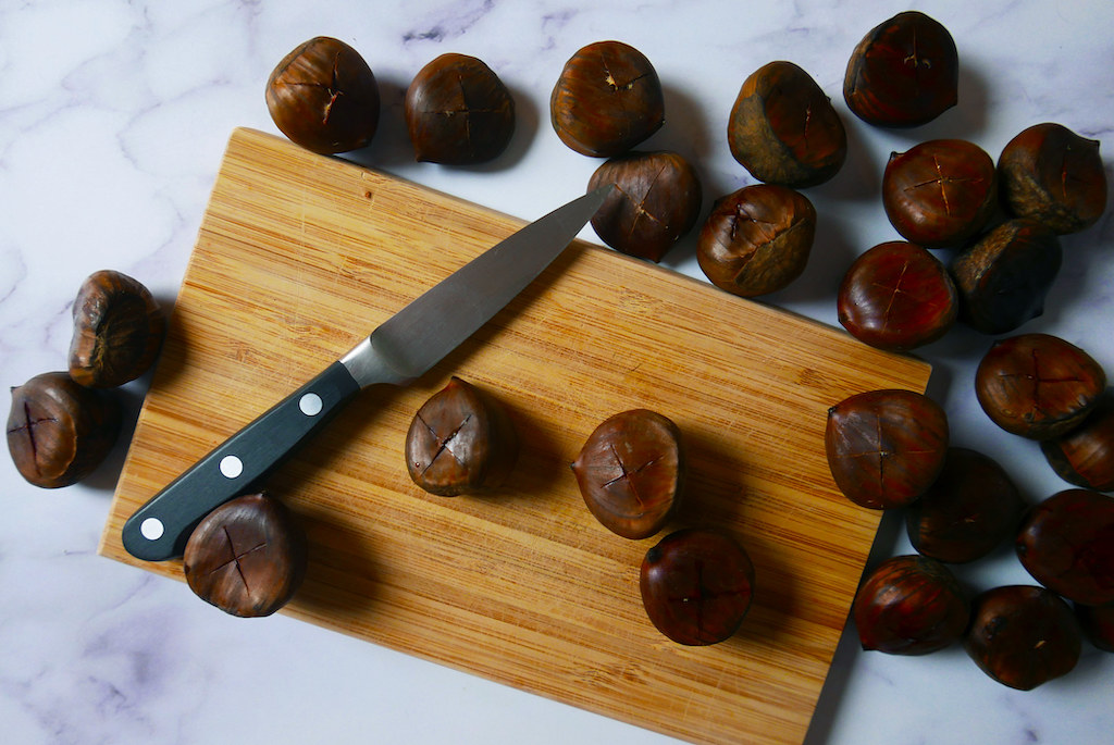 Chestnuts cut into an X pattern. Sitting on a cutting board with a pairing knife.