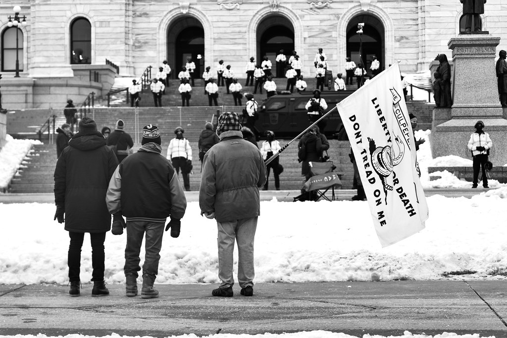A protester holding a Culpeper Minutemen flag at the Minnesota State Capitol in St Paul, Minnesota with state troopers on the Capitol steps.  Security is present at US state capitals across the country for possible violence in advance of President-elect J