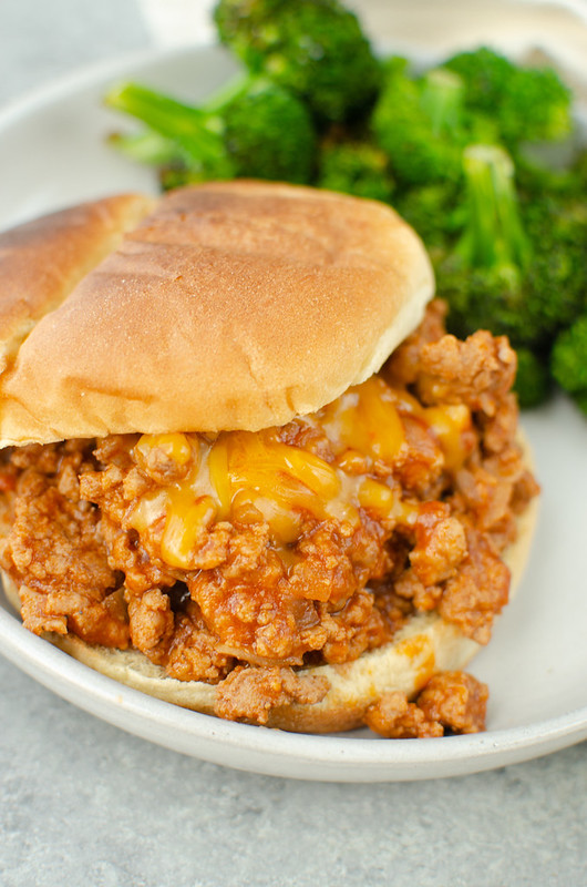Sloppy joe meat with shredded cheddar cheese on top, all on a toasted bun. With broccoli in the background. 