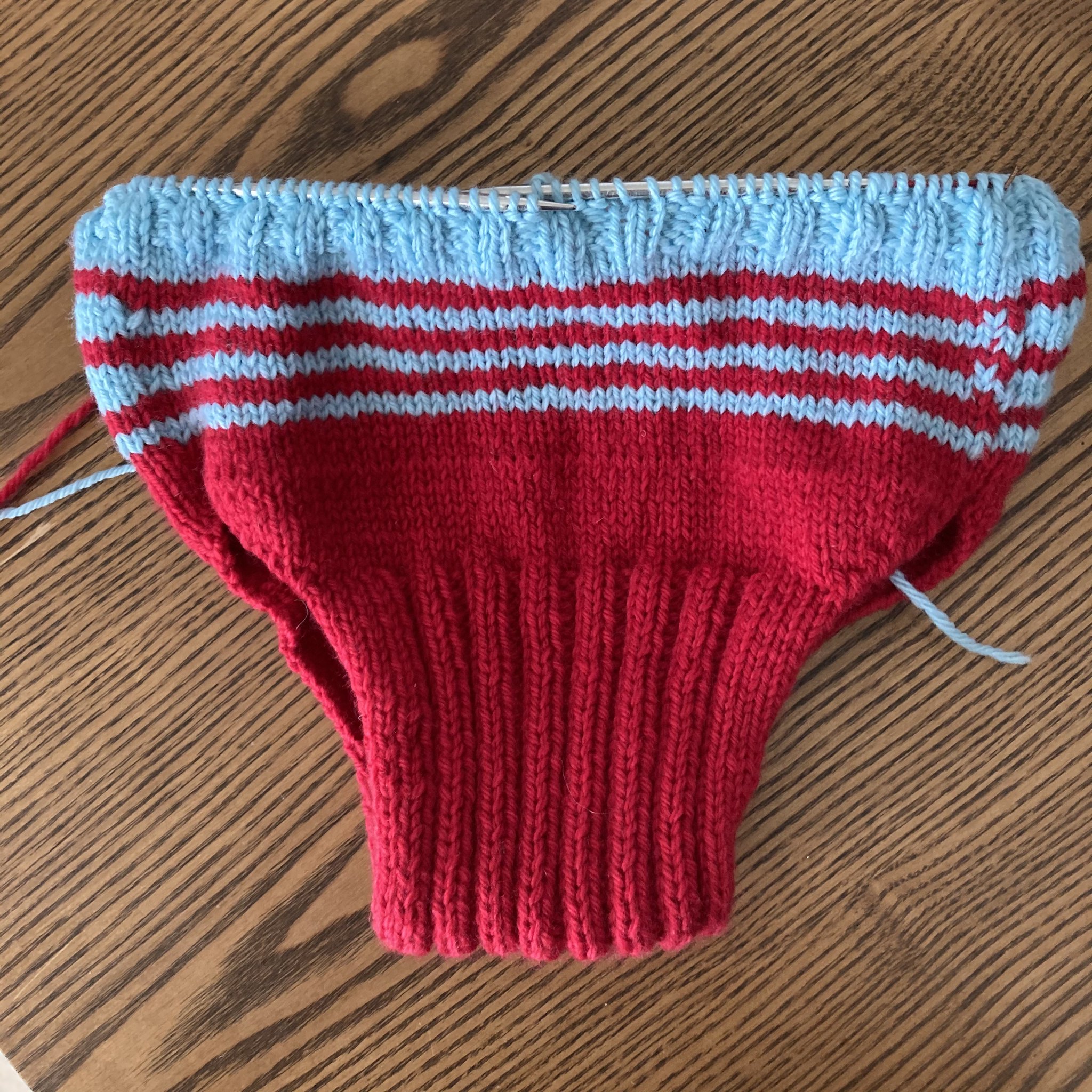 a knitted nappy cover, in progress. it is mainly red, with light blue stripes towards the top. the ribbing at the top has just begun