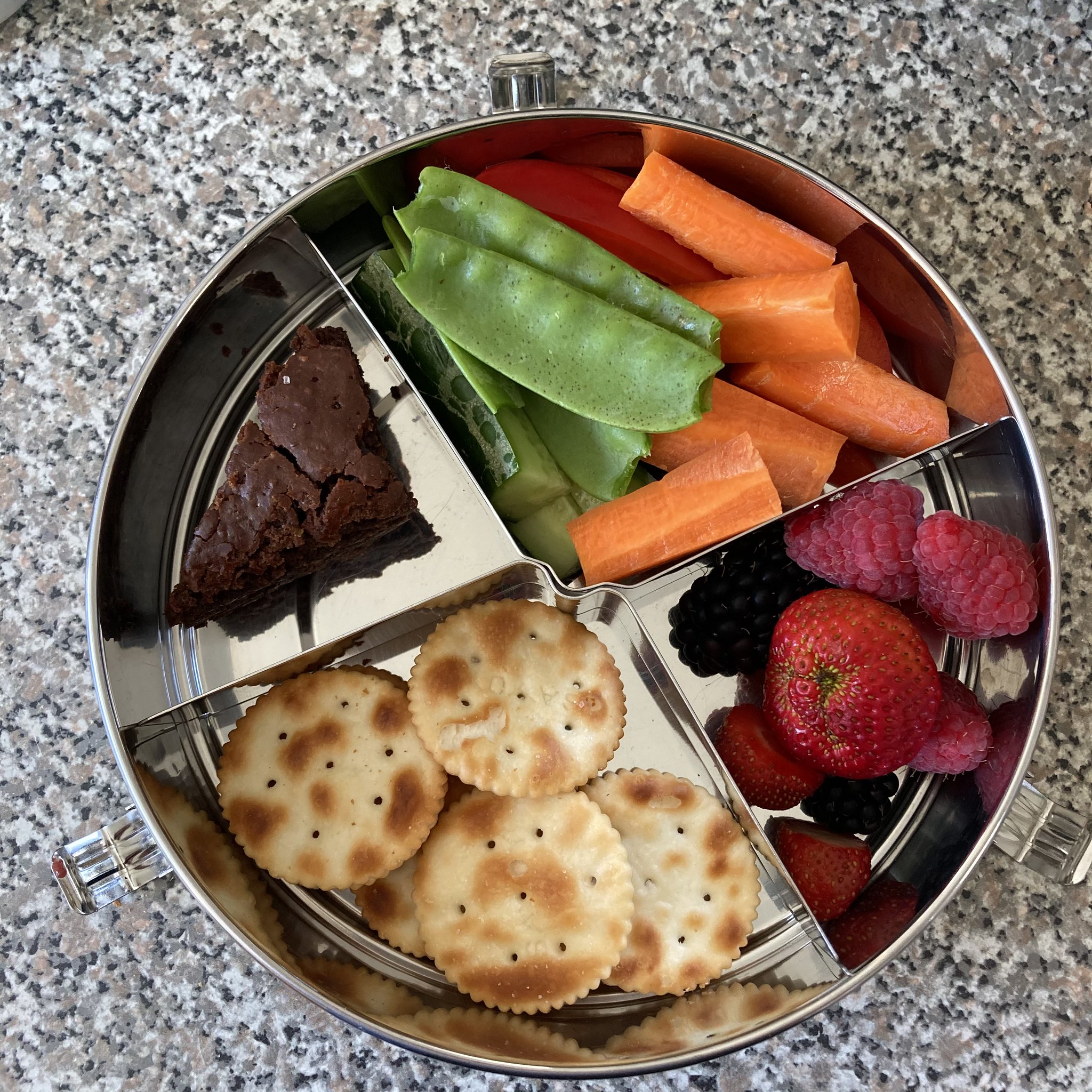 a round stainless steel lunchbox, packed with round crackers, a triangular piece of chocolate brownie, sticks of cucumber, capsicum, carrot and snowpeas and raspberries, blackberries and strawberries