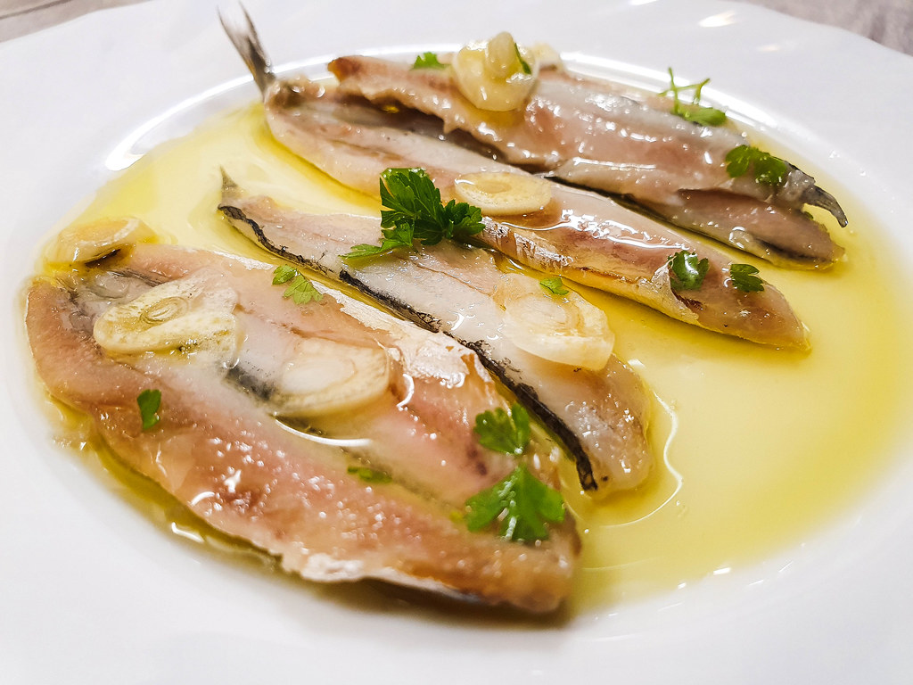 A white plate with six fillets of anchovies, covered with slices of garlic and chopped parsley, and drizzled with olive oil.