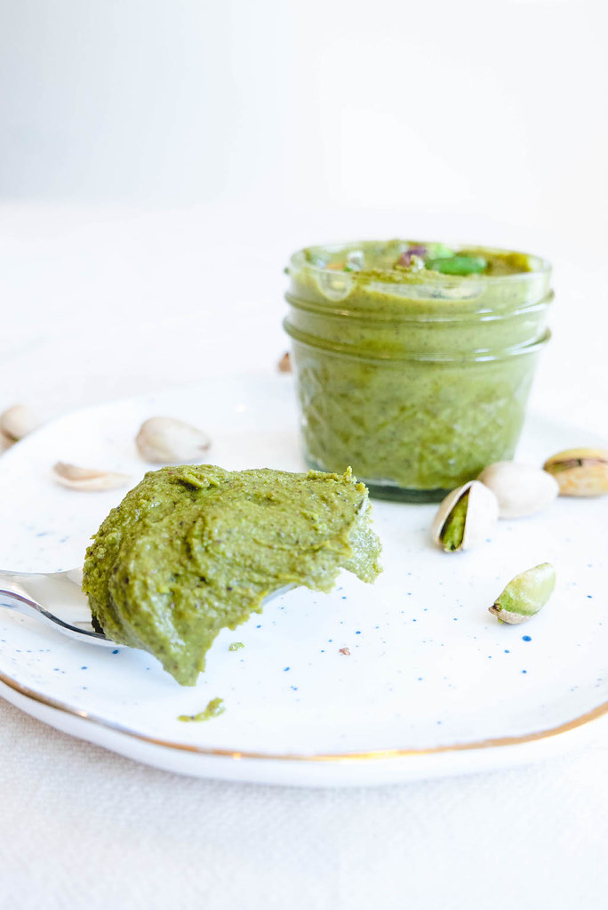 A spoonful of pistachio butter. The butter sits in a glass jar. 