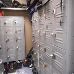 Photo shows a locker room with shoes lined up under them and bags and clothing piled on top.