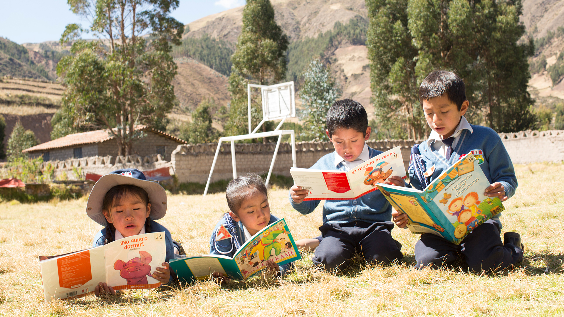 Four young children reading books on a patch of grass outside of a rural school building.