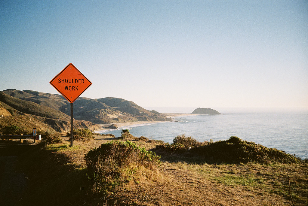 A hilly Pacific coastal scene, accompanied by a luminous orange road sign displaying 'SHOULDER WORK'