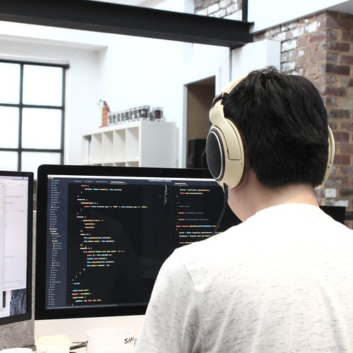 A person with headphones on, with their back turned away. They are working on their computer, which shows multiple lines of code. (Pre Apprenticeship Program)