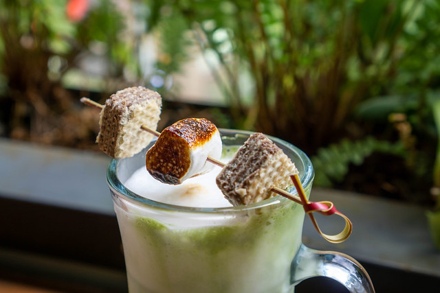 Geröstetes Marshmallow auf einem Holzspießchen mit Keksen auf einem Kaffeeglas mit Latte Macchiato Nahaufnahme