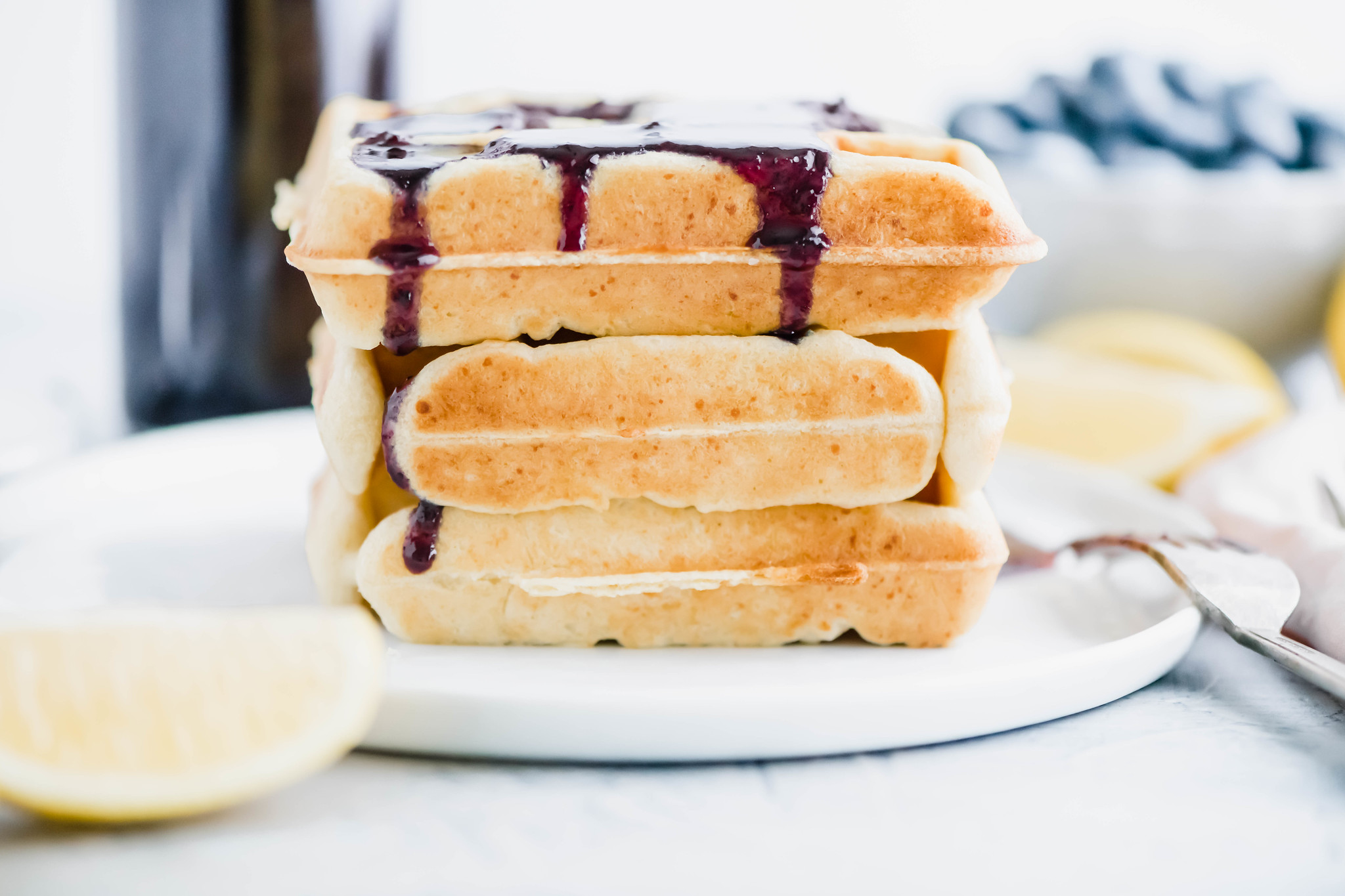 Three waffles stacked on white round plate with blueberry sryup dripping down front.