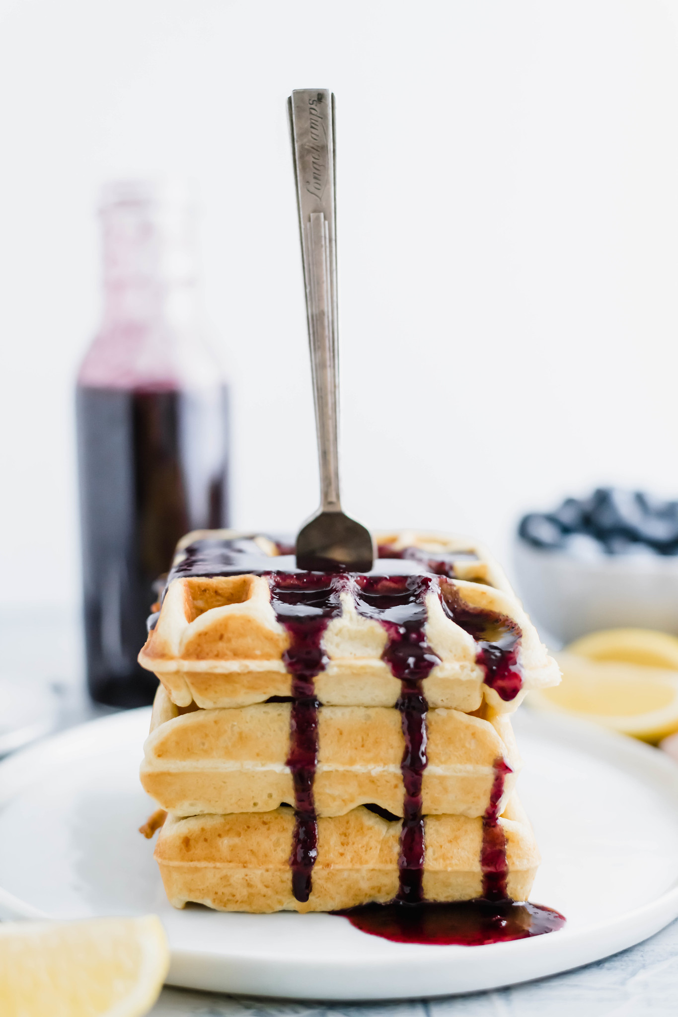 Three lemon ricotta waffles stacked on a white plate with blueberry sryup on top and dripping down. Fork stuck into the stack. Bottle of blueberry syrup in upper left corner. Bowl of blueberries in upper right corner.