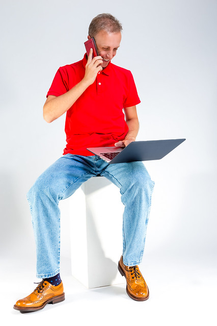 Distant Working Ideas. Portarit of Positive Caucasian Male in Red T-Shirt Posing With Laptop While Talking On Cellphone On White Background.