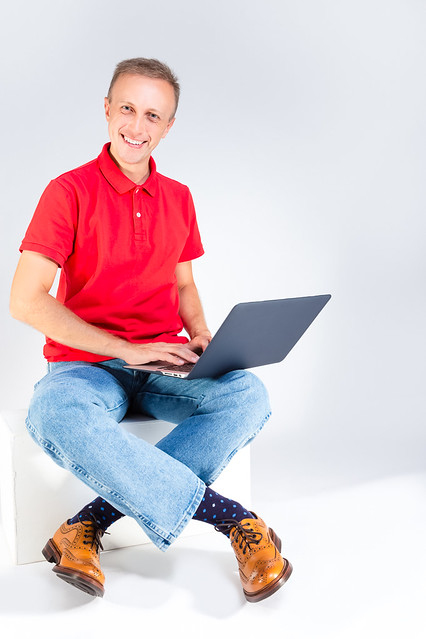 Distant Working Ideas. Full Length Portarit of Smiling Caucasian Handsome Man in Red T-Shirt Sitting With Laptop On White Background.