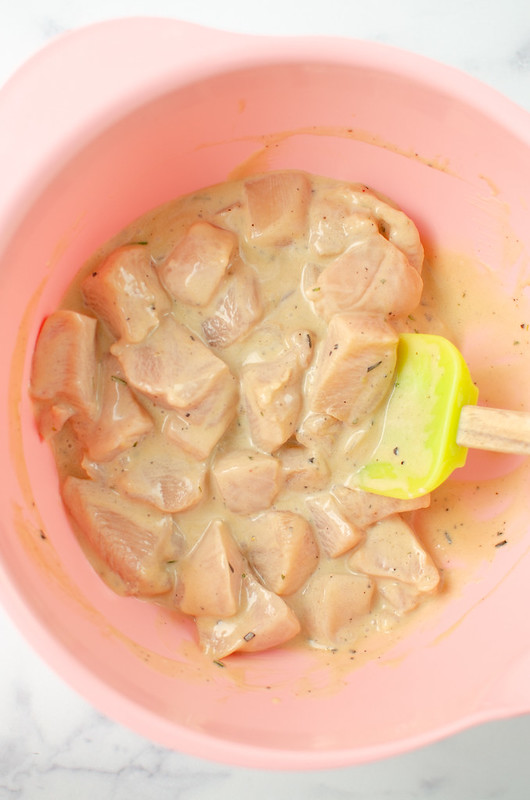 Chunks of chicken marinating in a rosemary and ranch marinade in a pink bowl with a yellow rubber spatula