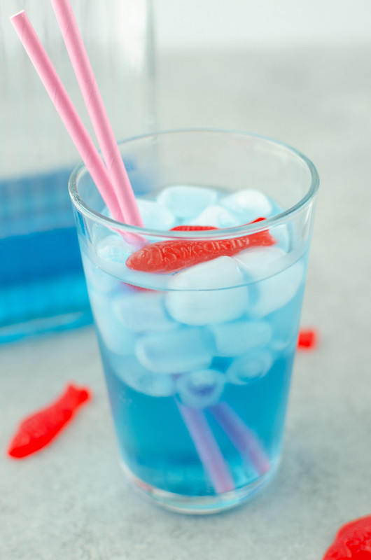blue soda, ice, and Swedish fish gummy candy in a glass; pitcher of soda in background and extra Swedish fish scattered around