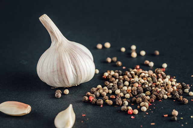 Various kinds of pepper and cloves of garlic next to a whole garlic head on black background