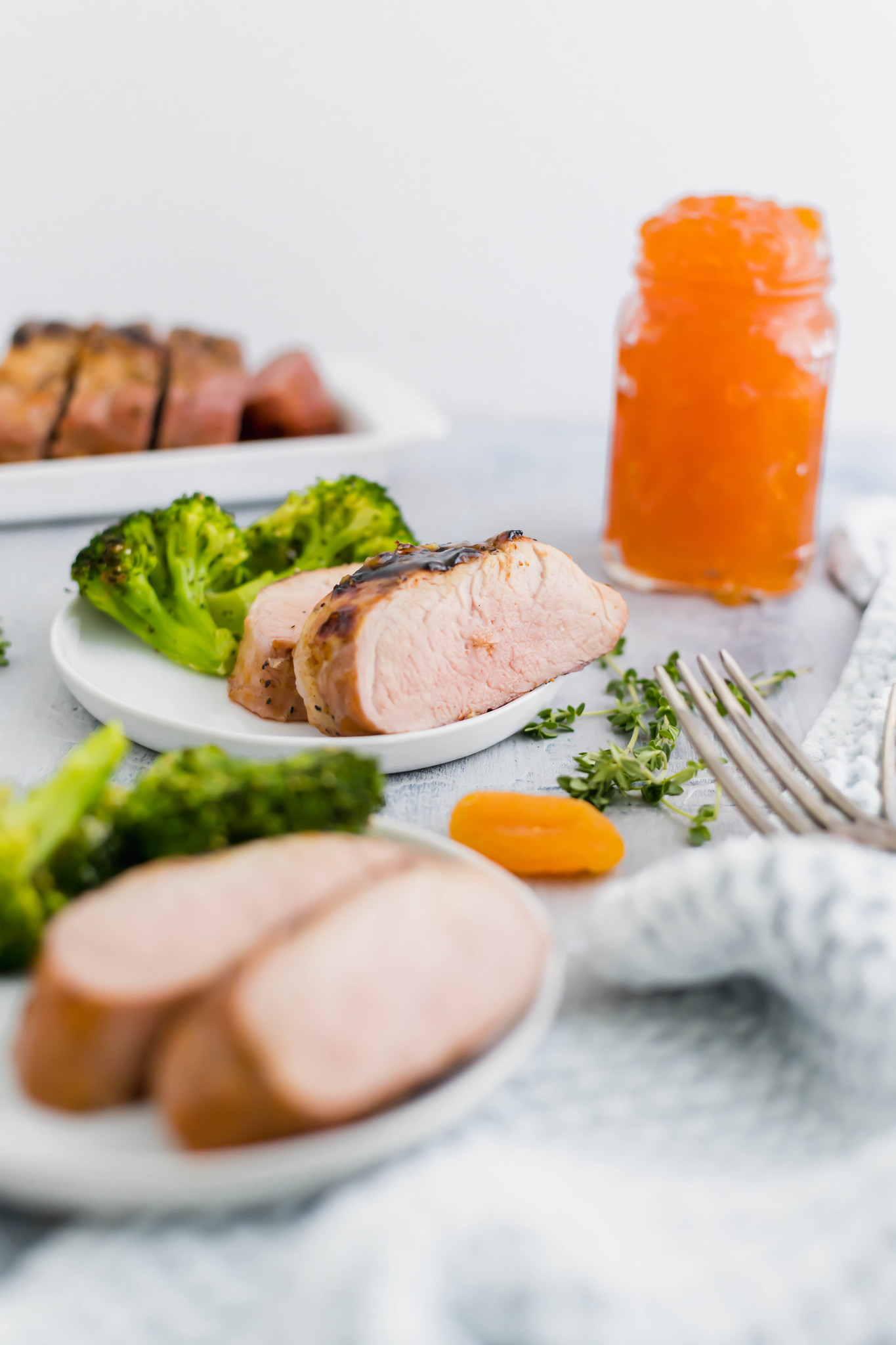 Two white round plates with two slices each of pork tenderloin and roasted broccoli. Plate in background in focus along with a jar of apricot preserves.