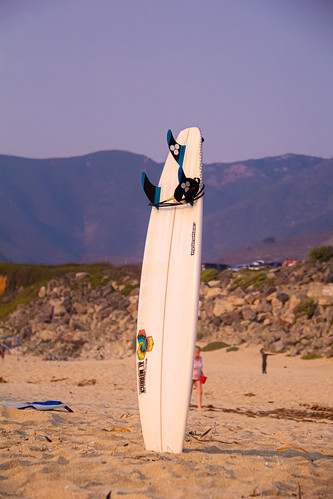 California, USA by Zⅰa, Surfboard propped up in the sand with rocky hills and colorful moutains for a backdrop. Taken with C