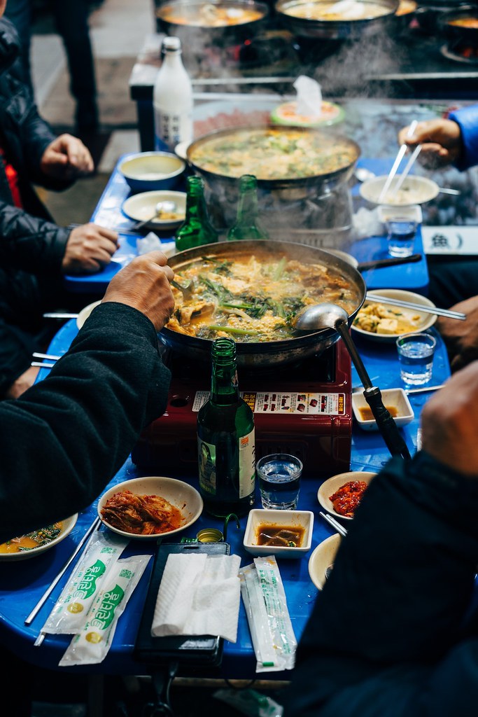 A shot of people eating stew on blue tables. Soju sits on the table next to side dishes. 