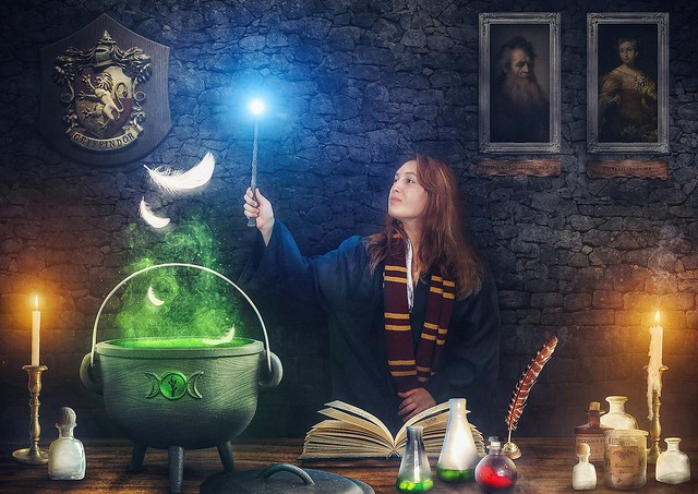 Harry Potter inspired photo compositing showing a women standing at a desk making potion. The desk is directly in front of a wall that has the crest of 'Gryffindor' on the left and two portraits of old wizards on the right. The woman is clothed in Gryffindor clothes (red &amp; yellow). A couple of items are placed on the desk: Candles, a large potion cauldron, an open book with instructions, and a couple of smaller test tubes