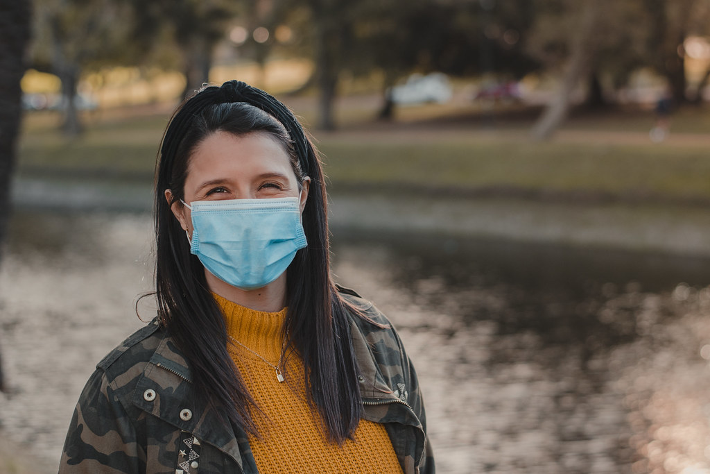 Horizontal shot of a Latina woman in a mask standing alone outdoors, looking at the camera and smiling. Outdoors concept in times of coronavirus. Copy space