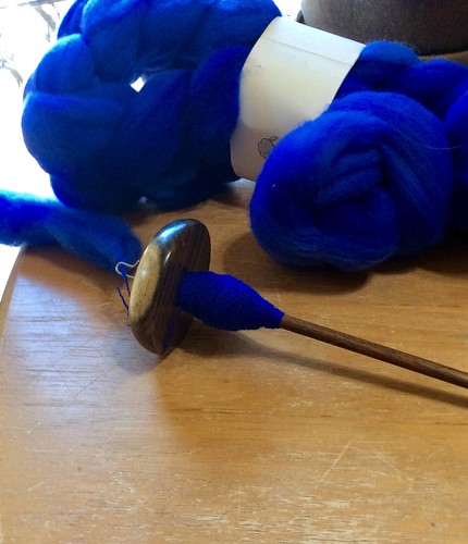 A braid of hand-dyed royal blue Targhee wool lies on a light wood table behind a Bosworth top-whorl drop spindle in Blue Mahoe wood with the start of spinning on the cop.  The spindle is laying horizontally in front of the horizontal braid of fibre and the hook, top-whorl are to the left with working fibre also to the left away from the whorl.