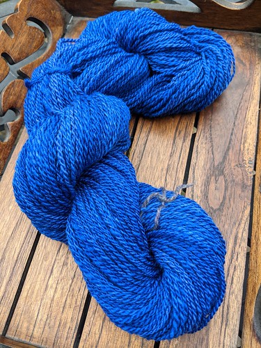 A skein of handspun royal blue Polwarth/silk yarn by irieknit handdyed by Sheepy Time Knits is tied and twisted on a small wood tray with a slatted bottom and gingerbread-style cut sides.