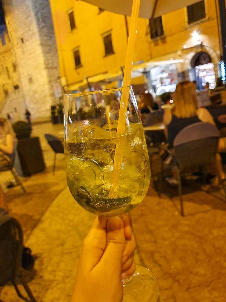 My hand holding a glass with a green drink in it. It is dark, and the photo is covered with a yellow light. In the background you can see people sitting at tables, blurred.