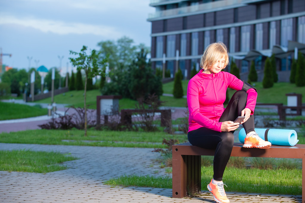 Blond Senior Female in Sport Outfit Posing With Smartphone While Resting With Travel Gear on Bench Outdoors.