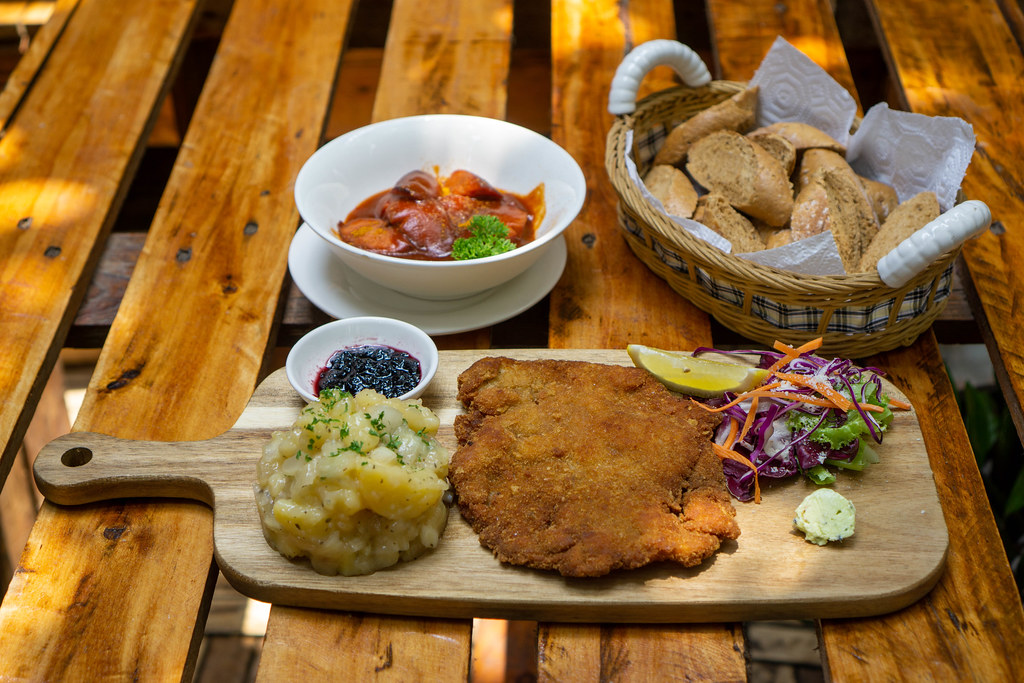 Food Photo of Chicken Schnitzel with Potato Salad on a Wooden Board, Currywurst in a Ceramic Bowl and a Basket of Bread Slices on a Wooden Table in a German Restaurant