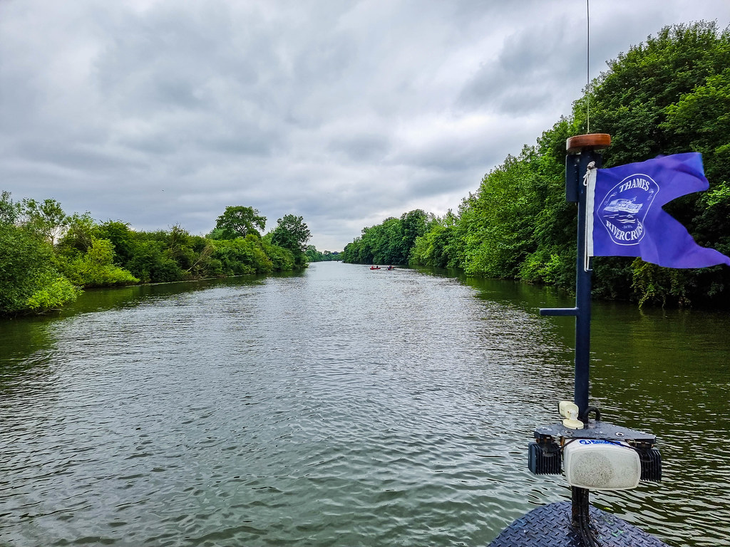 A view of the River Thames from the boat. There are green trees on both sides and the river is in the middle. In front you can see the blue flag of the boat company, Thames Rivercruise with its name written on it in white letters
