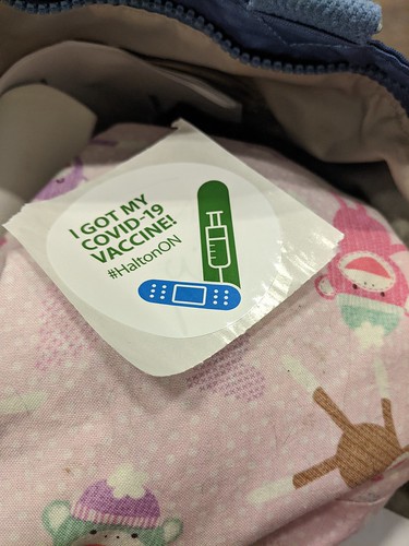 Sticker that reads I got my COVID-19 Vaccine hashtag HaltonON in green text on white background above a blue stylized band-aid and a green stylized syringe rests on a cloth drawstring bag with pink background, white polka dots and knit sock monkey motif inside a larger bag.